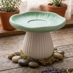 May include: A ceramic mushroom-shaped pedestal stand featuring a pale green cap with a smooth, rounded edge and a white, vertically ribbed stem. The base of the stem is surrounded by a small cluster of natural green moss and smooth gray river stones.