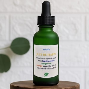 Könnte beinhalten: Eine grüne Glasflasche mit schwarzer Pipette, beschriftet mit "JUST BE HAPPY" mit ätherischen Ölen. Die Flasche steht auf einer kleinen, runden Holzkiste. Der Hintergrund ist eine weiße Ziegelmauer.