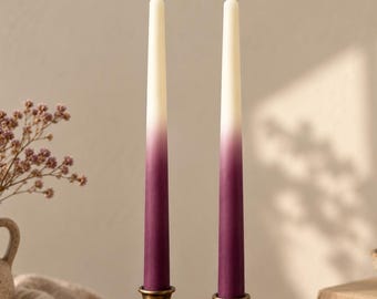 Op de afbeelding: Twee hoge taps toelopende kaarsen met een gladde ombre-gradiënt die overgaat van een dieppaarse basis naar een romige gebroken witte top. Elke kaars wordt rechtop gehouden door een klassieke koperen kandelaar met een brede, ronde voet.