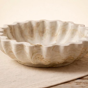 Op de afbeelding: Een decoratieve, gebroken witte marmeren kom met een geschulpte rand. De kom heeft een natuurlijk, wervelend patroon van beige en crèmetinten. Hij staat op een neutraal gekleurd linnen oppervlak, geschikt voor een keuken of eetkamer.