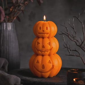 May include: A stack of three orange wax pumpkins shaped into a candle with a lit wick at the top. Each pumpkin features a carved face with cutouts for eyes, a nose, and a mouth. The top pumpkin has a simple face, while the middle and bottom pumpkins display wider, jagged grins.