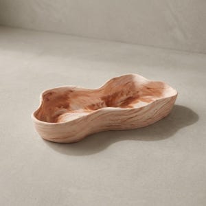 May include: A light brown, oblong-shaped ceramic bowl with a marbled effect. The bowl has a wavy rim and a shallow interior. The color palette includes shades of light and dark brown, creating a natural, organic aesthetic. Ideal for serving snacks or as a decorative piece.