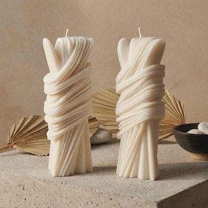 Può includere: Due candele scultoree color avorio. Ogni candela presenta un design unico con cera a spirale e stratificata, creando un effetto drappeggiato strutturato. Le candele sono in posizione verticale, con uno stoppino in cima. Lo sfondo include foglie di palma secche e una piccola ciotola.