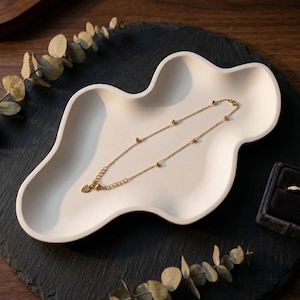 May include: A gold-colored necklace with a heart pendant and small round beads rests on a white, cloud-shaped tray. The tray sits on a dark, textured surface, with a small ring box visible in the background. Eucalyptus leaves add a natural touch.