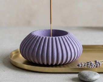 May include: A lavender-colored incense holder with a ribbed design, holding a lit incense stick. The holder sits on a gold-colored tray, with a small stone and sprig of lavender nearby. The overall theme is home decor and aromatherapy.