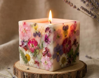 Peut inclure: Une bougie carrée allumée ornée de fleurs séchées roses, violettes, jaunes et blanches. La bougie repose sur une petite base en bois ronde. L'arrière-plan est un tissu neutre.
