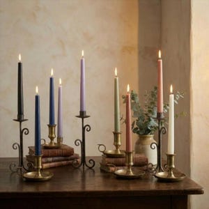 Puede incluir: Una colección de velas cónicas encendidas en varios colores, incluyendo negro, azul, morado, verde, rosa y crema. Las velas están en candelabros ornamentados de metal y latón, dispuestas sobre una superficie de madera oscura. Algunas están sobre libros.