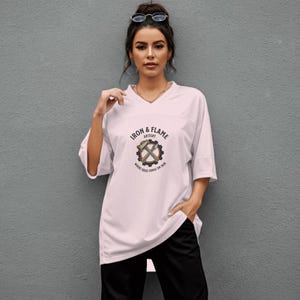 K&ouml;nnte beinhalten: Wei&szlig;es kurz&auml;rmeliges Trikot mit V-Ausschnitt, mit dem Text "IRON & FLAME ARTISTRY" und einer kreisf&ouml;rmigen Grafik mit Werkzeugen. Das Trikot wird mit schwarzen Sporthosen getragen.