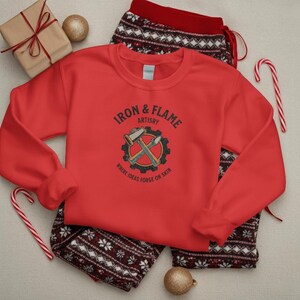 K&ouml;nnte beinhalten: Ein rotes Sweatshirt mit dem Text "IRON & FLAME ARTISTRY" und einer Grafik von Hammer und Mei&szlig;el. Das Sweatshirt wird mit einer gemusterten Pyjamahose, einem Geschenk und Zuckerstangen gezeigt.