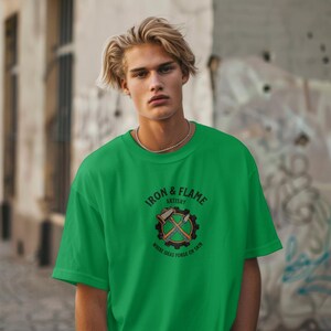 K&ouml;nnte beinhalten: Ein leuchtend gr&uuml;nes T-Shirt mit dem Text "IRON & FLAME ARTISRY" und einer Grafik von gekreuzten Werkzeugen in einem Zahnrad. Das Rundhals-Shirt zeigt den Slogan "WHERE IDEAS FORGE ON SKIN" unter dem zentralen Design. Das T-Shirt ist ein kr&auml;ftiges Gr&uuml;n.
