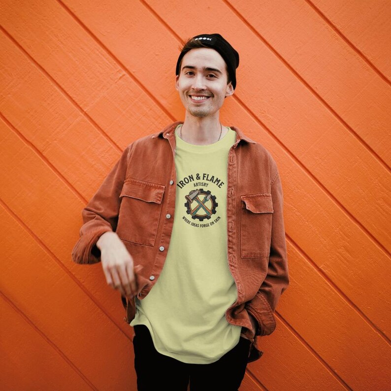 K&ouml;nnte beinhalten: Eine Person tr&auml;gt ein hellgelbes T-Shirt mit dem Text "IRON & FLAME ARTISTRY" und einer Zahnrad- und gekreuzten &Auml;xte-Grafik. Die Person tr&auml;gt auch eine braune Jacke und eine schwarze M&uuml;tze.