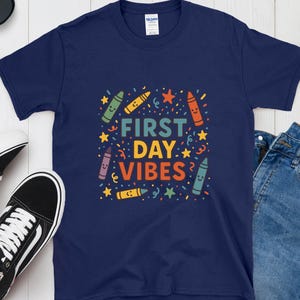 Könnte beinhalten: Marineblaues T-Shirt mit dem Schriftzug "FIRST DAY VIBES" in bunten Farben. Das Design umfasst lächelnde Buntstifte, Sterne und Konfetti. Ein fröhliches Design für den Schulanfang.