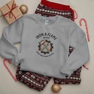 K&ouml;nnte beinhalten: Ein hellgraues Sweatshirt mit dem Text "Iron & Flame Artistry" und einer Grafik eines Hammers und eines Mei&szlig;els in einem Zahnrad. Das Sweatshirt liegt auf einer gemusterten Pyjamahose, mit einem verpackten Geschenk, Ornamenten und Zuckerstangen.