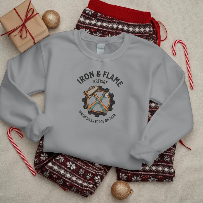 K&ouml;nnte beinhalten: Ein hellgraues Sweatshirt mit dem Text "Iron & Flame Artistry" und einer Grafik eines Hammers und eines Mei&szlig;els in einem Zahnrad. Das Sweatshirt liegt auf einer gemusterten Pyjamahose, mit einem verpackten Geschenk, Ornamenten und Zuckerstangen.