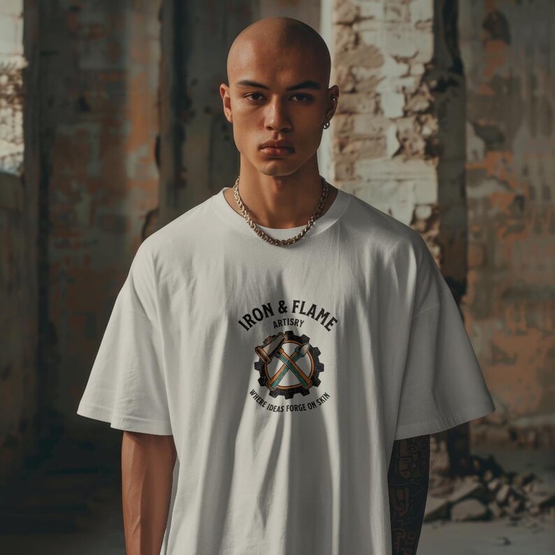 K&ouml;nnte beinhalten: Wei&szlig;es &uuml;bergro&szlig;es T-Shirt mit dem Text "IRON & FLAME ARTISRY" und einer Grafik eines Zahnrads, Hammers und gekreuzten &Auml;xten. Das Shirt hat einen Rundhalsausschnitt und kurze &Auml;rmel. Das Model tr&auml;gt eine silberne Halskette.