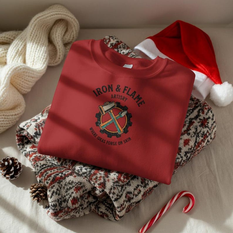 K&ouml;nnte beinhalten: Ein rotes Sweatshirt mit dem Text "IRON & FLAME ARTISTRY" und einer Grafik von Hammer und Mei&szlig;el in einem Zahnrad. Das Sweatshirt liegt gefaltet auf einem gemusterten Pullover, mit einem Weihnachtsmannhut und einer Zuckerstange im Hintergrund.