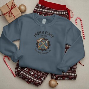 K&ouml;nnte beinhalten: Ein schieferblaues Sweatshirt mit dem Text "IRON & FLAME ARTISTRY" und einer Grafik von Hammer und Mei&szlig;el. Das Sweatshirt liegt auf einer rot-wei&szlig; gemusterten Pyjamahose. Ein verpacktes Geschenk, eine goldene Kugel und Zuckerstangen sind ebenfalls im Bild.