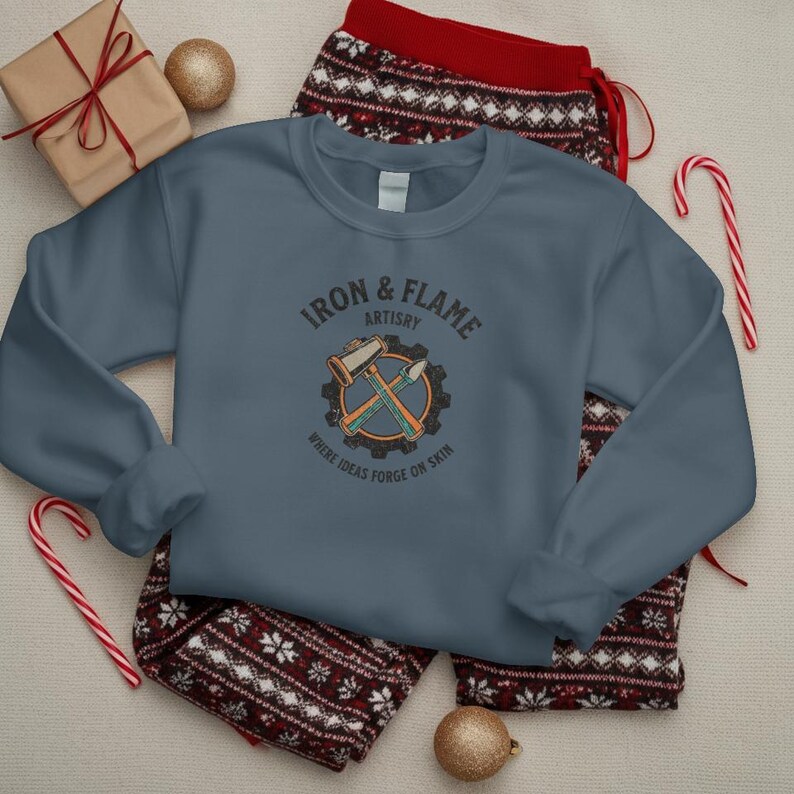 K&ouml;nnte beinhalten: Ein schieferblaues Sweatshirt mit dem Text "IRON & FLAME ARTISTRY" und einer Grafik von Hammer und Mei&szlig;el. Das Sweatshirt liegt auf einer rot-wei&szlig; gemusterten Pyjamahose. Ein verpacktes Geschenk, eine goldene Kugel und Zuckerstangen sind ebenfalls im Bild.