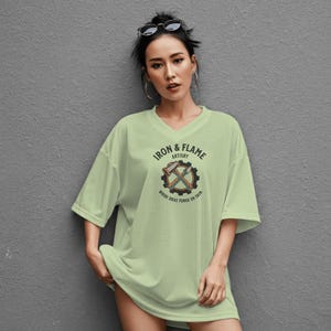 K&ouml;nnte beinhalten: Ein hellgr&uuml;nes, &uuml;bergro&szlig;es V-Ausschnitt-T-Shirt mit einem Aufdruck auf der Vorderseite. Der Aufdruck zeigt den Text "IRON & FLAME ARTISTRY" um ein Zahnrad mit gekreuzten H&auml;mmern und &Auml;xten. Der Text "WHERE IDEAS FORGE ON SKIN" steht darunter.