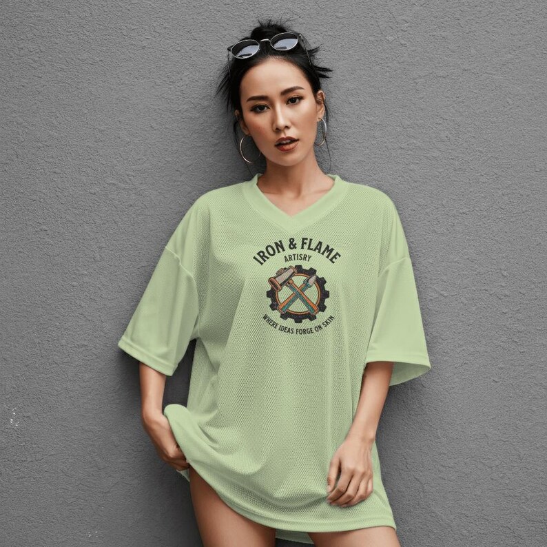 K&ouml;nnte beinhalten: Ein hellgr&uuml;nes, &uuml;bergro&szlig;es V-Ausschnitt-T-Shirt mit einem Aufdruck auf der Vorderseite. Der Aufdruck zeigt den Text "IRON & FLAME ARTISTRY" um ein Zahnrad mit gekreuzten H&auml;mmern und &Auml;xten. Der Text "WHERE IDEAS FORGE ON SKIN" steht darunter.