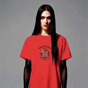 K&ouml;nnte beinhalten: Ein rotes T-Shirt mit dem Text "IRON & FLAME ARTISTRY" und einer Grafik von gekreuzten Werkzeugen in einem Zahnrad. Das T-Shirt ist kurz&auml;rmlig und hat einen Rundhalsausschnitt. Das Model hat lange schwarze Haare.