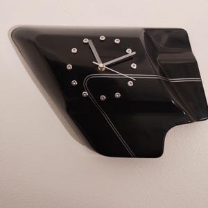 May include: A black motorcycle part clock with silver hands and white accents. The clock face has a unique design with a black background and white lines.