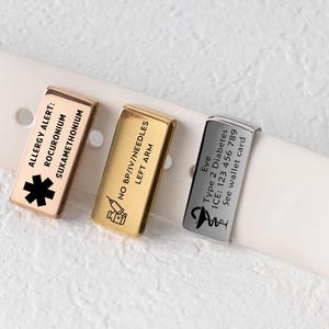 May include: Three medical alert charms in gold, silver, and rose gold colors, attached to a white watch band. Each charm has engraved text, including allergy and medical information. The charms are rectangular with rounded edges.