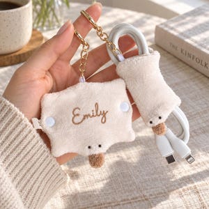 May include: A cream-coloured plush cable organiser featuring a gold-tone metal keychain. The rectangular fabric piece is embroidered with the name Emily in brown script, includes two white snap buttons, and has a small brown plush nose detail.