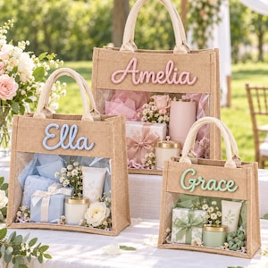 May include: Three natural jute tote bags with clear plastic windows and fabric handles. Each bag features a personalized name in raised script: Amelia in pink, Ella in blue, and Grace in green. The bags are filled with gift items including candles, flowers, and wrapped boxes.