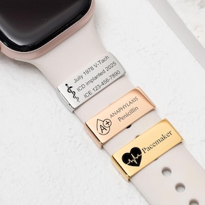 Puede incluir: Primer plano de un smartwatch con una correa rosa claro, que muestra tres dijes de alerta médica. Los dijes son plateados, oro rosa y dorados, cada uno grabado con información y símbolos médicos.