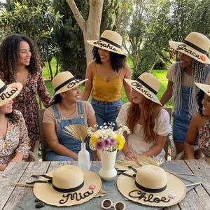 May include: A group of women wearing personalised straw hats with black bands and names embroidered on the brims. The hats are beige with black lettering. The women are sitting around a wooden table outdoors.