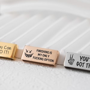 Puede incluir: Tres barras metálicas rectangulares con frases motivadoras. La primera es dorada con un pato con gafas de sol y el texto "You Can DO IT!". La segunda es oro rosa con una cara aterradora y el texto "Finishing is my only fucking option". La tercera es plateada con una mano de esqueleto y el texto "You've Got This!"