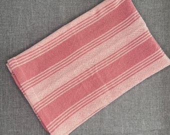 Puede incluir: Un textil rectangular plegado, con rayas rosas y blancas. El textil tiene rayas rosas y blancas alternas, creando una apariencia texturizada. La tela está hecha de material tejido. El textil se muestra sobre una superficie gris.