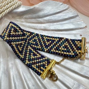 Puede incluir: Una pulsera de cuentas con un patrón geométrico en oro, azul y morado. La pulsera tiene cierres dorados y una cadena. La pulsera se muestra sobre una superficie blanca en forma de concha.