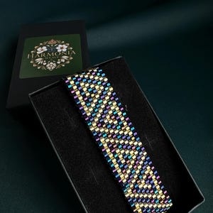 May include: A rectangular bracelet crafted from small, metallic beads in alternating rows of gold, purple, and teal. The bracelet is displayed inside a black box, which sits next to a black box lid featuring a green label with the text HARMONIA WEAVE.