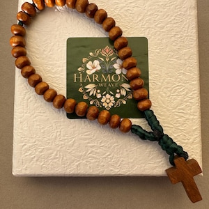 Può includere: Un rosario di legno con perline marroni e un cordoncino verde. Il rosario è in una scatola bianca con un'etichetta verde che dice "Harmon Weave".
