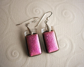May include: A pair of rectangular drop earrings featuring a vibrant, textured pink dichroic glass center layered over a dark base, topped with a clear, patterned glass section. The earrings are suspended from silver-toned metal ear wires.