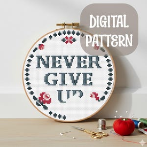 May include: A cross-stitch design in a wooden hoop, featuring the text "NEVER GIVE UP" in dark blue. The design includes red and white floral accents and a dotted border. The text "DIGITAL PATTERN" is in a grey bubble.