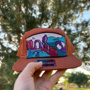 May include: A rust-colored, five-panel hat with a flat brim. The front panel features an embroidered landscape design with purple, blue, and pink elements. The hat is held by a hand, and a tag is visible below the brim.