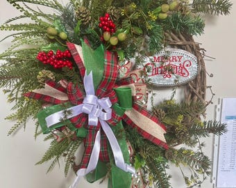 May include: A Christmas wreath with a rustic, natural design. It features a woven base adorned with evergreen branches, red berries, pinecones, and a decorative bow with red, green, and white ribbons. A metal sign reads "Merry Christmas."