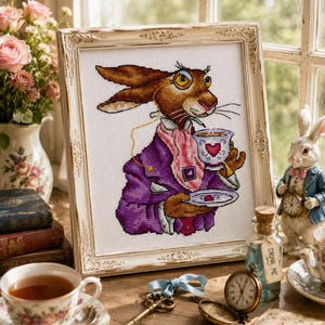 Könnte beinhalten: Eine gerahmte Kreuzstichstickerei, die einen braunen Hasen mit einer violetten Jacke, einem rosa Halstuch und einem Monokel darstellt, der eine Teetasse und eine Untertasse hält. Der Hase hat lange Ohren und ist detailliert gestickt.
