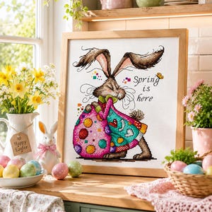 Puede incluir: Un bordado en punto de cruz enmarcado que muestra un conejo marrón de orejas largas sosteniendo un huevo grande con lunares de colores. El diseño incluye el texto "Spring is here" en letra negra, acentuado por una pequeña mariposa amarilla y cuadrados pixelados de colores.