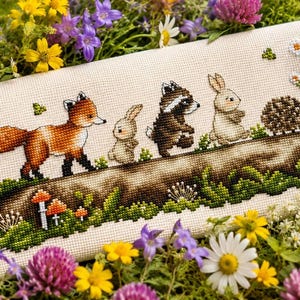 May include: A cross-stitch embroidery on light beige fabric featuring a horizontal log with a red fox, a small rabbit, a raccoon, another rabbit, and a hedgehog walking across it. The scene includes green moss, orange mushrooms, and white flowers.