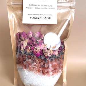 May include: A clear, resealable bag filled with botanical bath salts. The salts are layered with pink rose buds, lavender, and coarse salt crystals. A white label reads "DREAM" and "SOMA & SAGE". A small tea light candle sits on top.