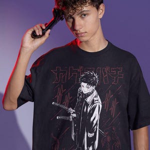 May include: Black t-shirt featuring a Japanese anime-style illustration. The graphic includes a character with swords and Japanese text in red. The shirt is worn by a person holding a black object.