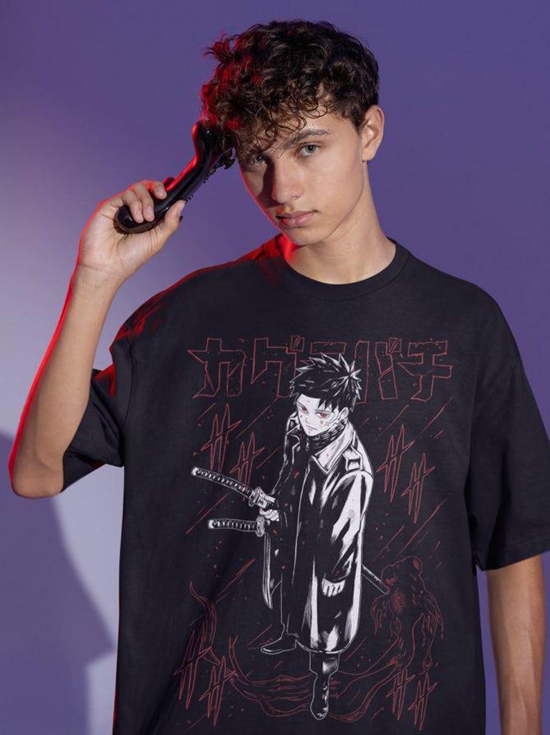 May include: Black t-shirt featuring a Japanese anime-style illustration. The graphic includes a character with swords and Japanese text in red. The shirt is worn by a person holding a black object.