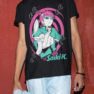 May include: Black t-shirt featuring a graphic of an anime character with pink hair and green glasses, holding a spoon. The design includes pink and white swirls and Japanese text. The text "Saiki K." is also visible.