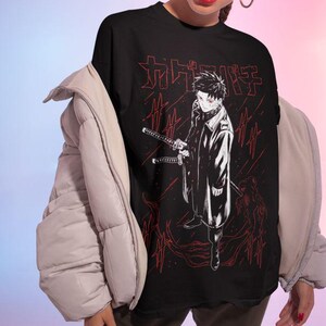 May include: Black t-shirt featuring a detailed anime-style illustration of a character holding two swords, with red Japanese text and accents. The design is set against a backdrop of red lines. The shirt is worn by a person.