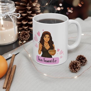 May include: White ceramic mug featuring a cartoon illustration of a woman holding a bowl and spoon, with the text "Fan Favorite" on a pink banner. The mug is filled with a dark liquid.