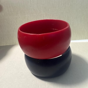 May include: A stack of two wide, smooth bangle bracelets. The top bracelet is a solid, vibrant red color, while the bottom bracelet is a solid, deep black color. Both bracelets feature a rounded, thick profile with a simple, unadorned design.